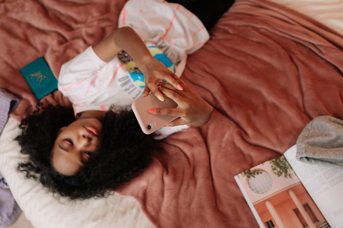 A teenage girl lies on her bed holding a smartphone above her face