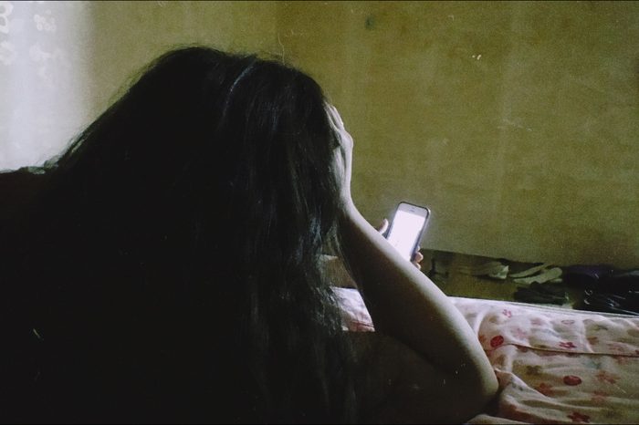 A young person lies awake in a dim bedroom, face turned toward a glowing phone screen